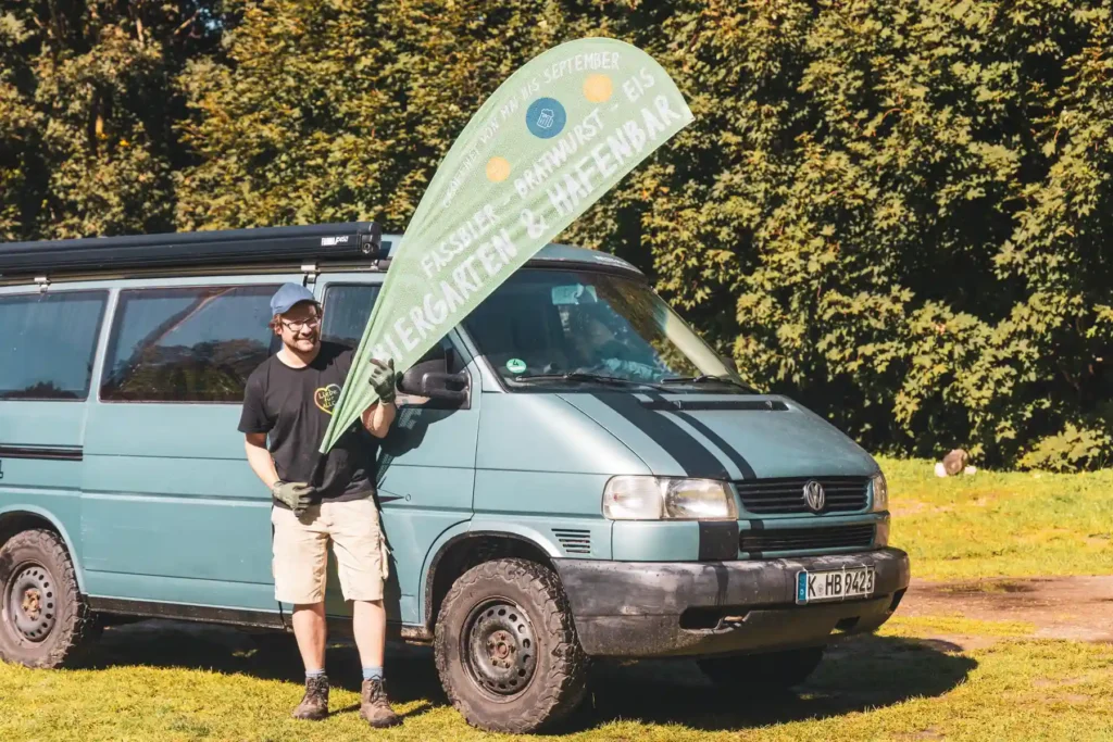 Naturcamping Bermudadreieck Geheimtipp Mecklenburgische Seenplatte Naturcampingplatz Bermudadreieck Betreiber Rainer mit Campingplatzflagge vor VW Bus Camper